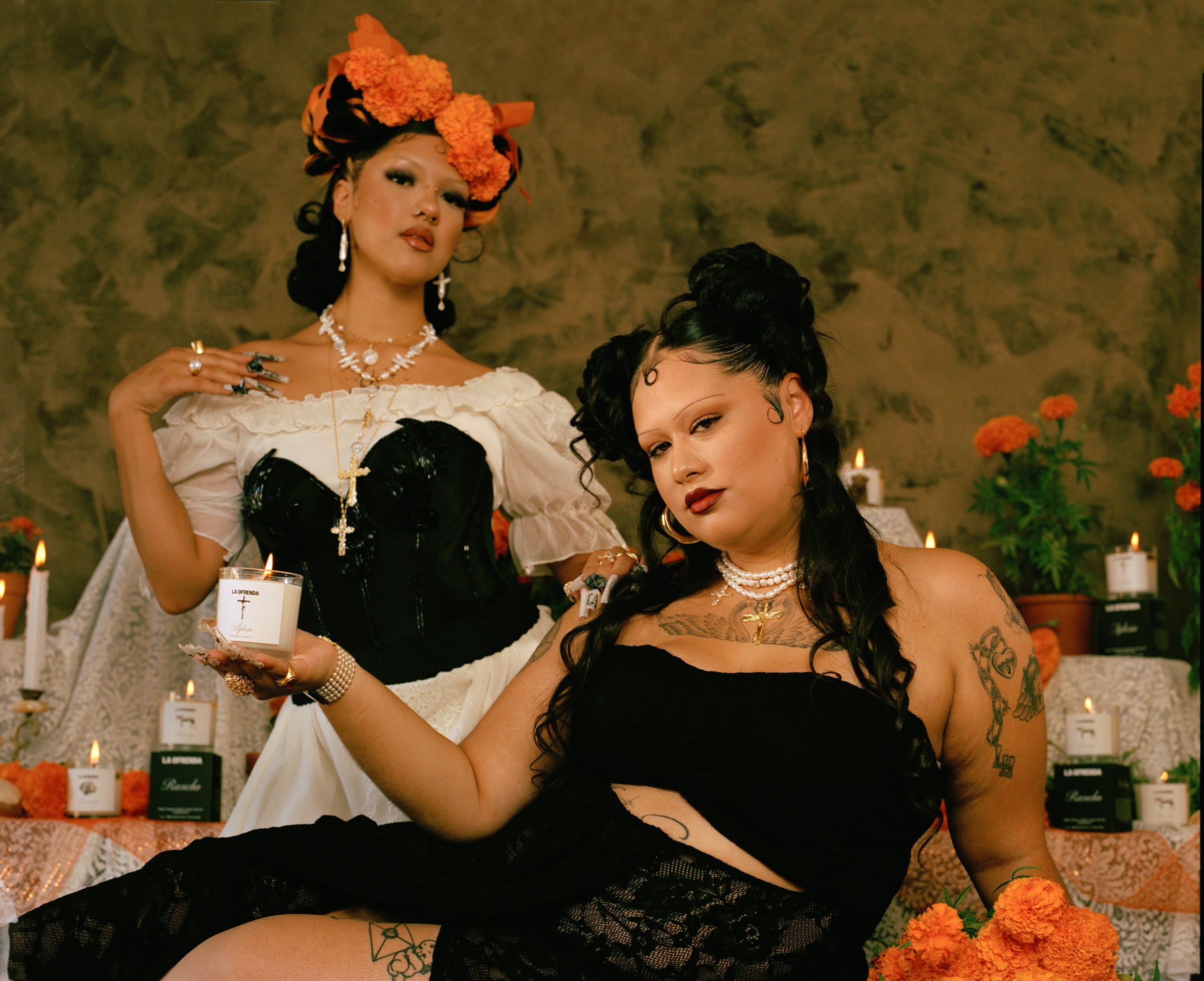 Two women in elaborate costumes with floral headpieces and jewelry, sitting in a decorated setting with candles and flowers.